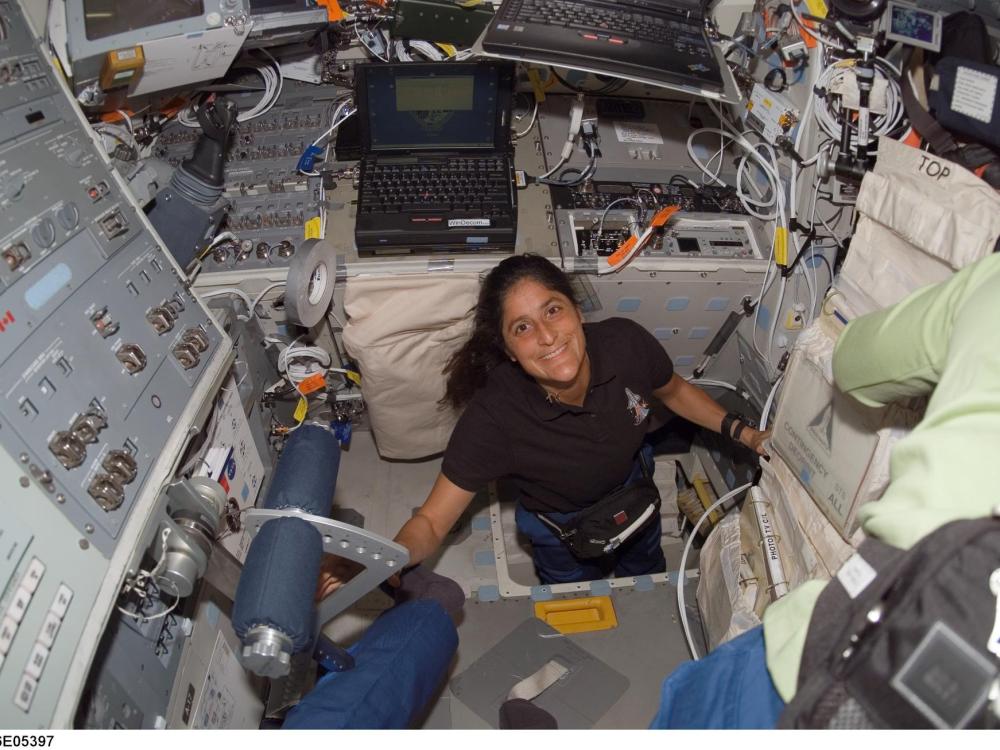 Sunita Williams, an astronaut, emerges through a hatch on the Space Shuttle into a room full of technical equipment. She's smiling. 