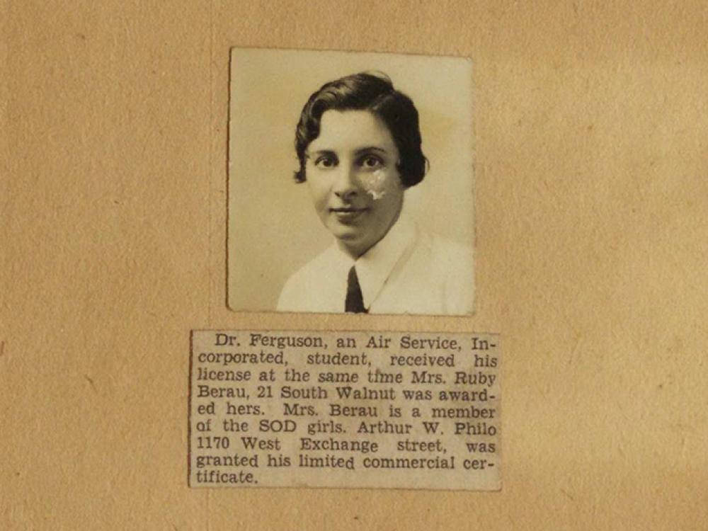 A close snapshot of Rubye Berau's yellowing scrapbook shows her picture and a newspaper clipping that reads "Dr. Ferguson, an Air Service Incorporated student received his license at the same time Mrs. Ruby Berau, 21 South Walnut was awarded hers. Mrs. Berau is a member of the SOD girls. Arthur W. Philo granted his limited commercial certificate."