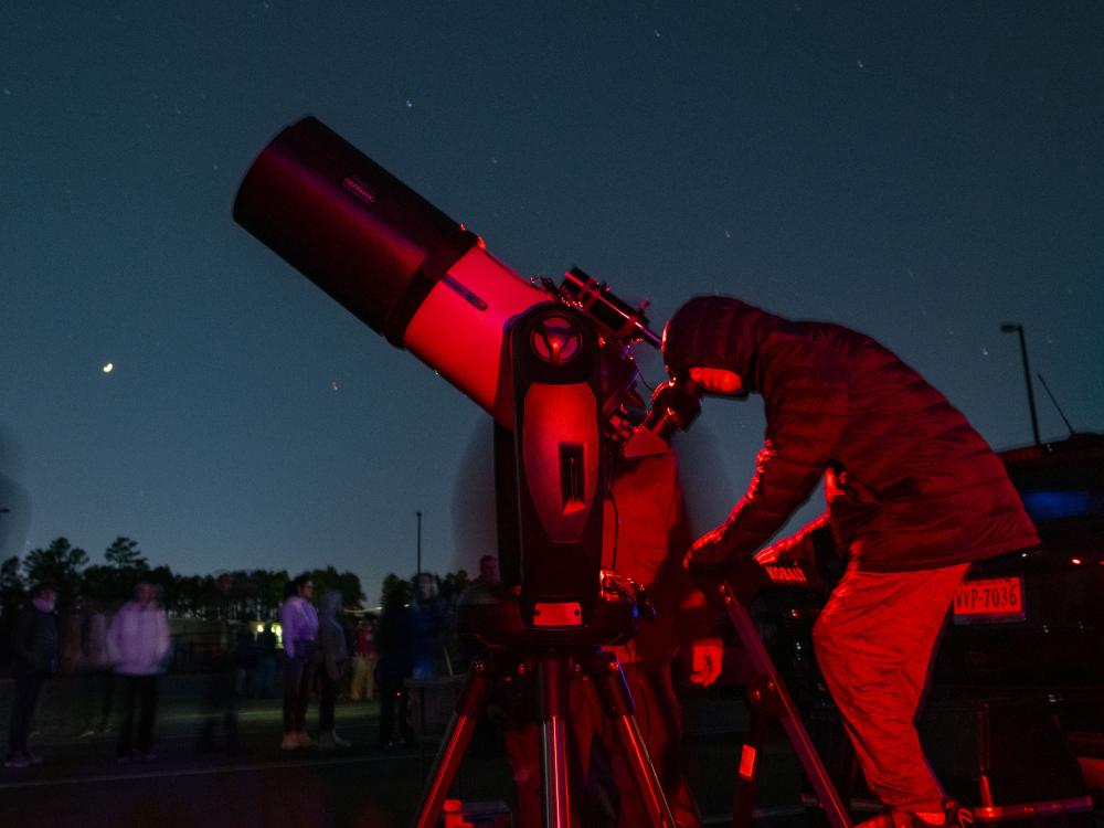 A young person stands on a stepladder and looks through a large telescope at a dark blue-green night sky.