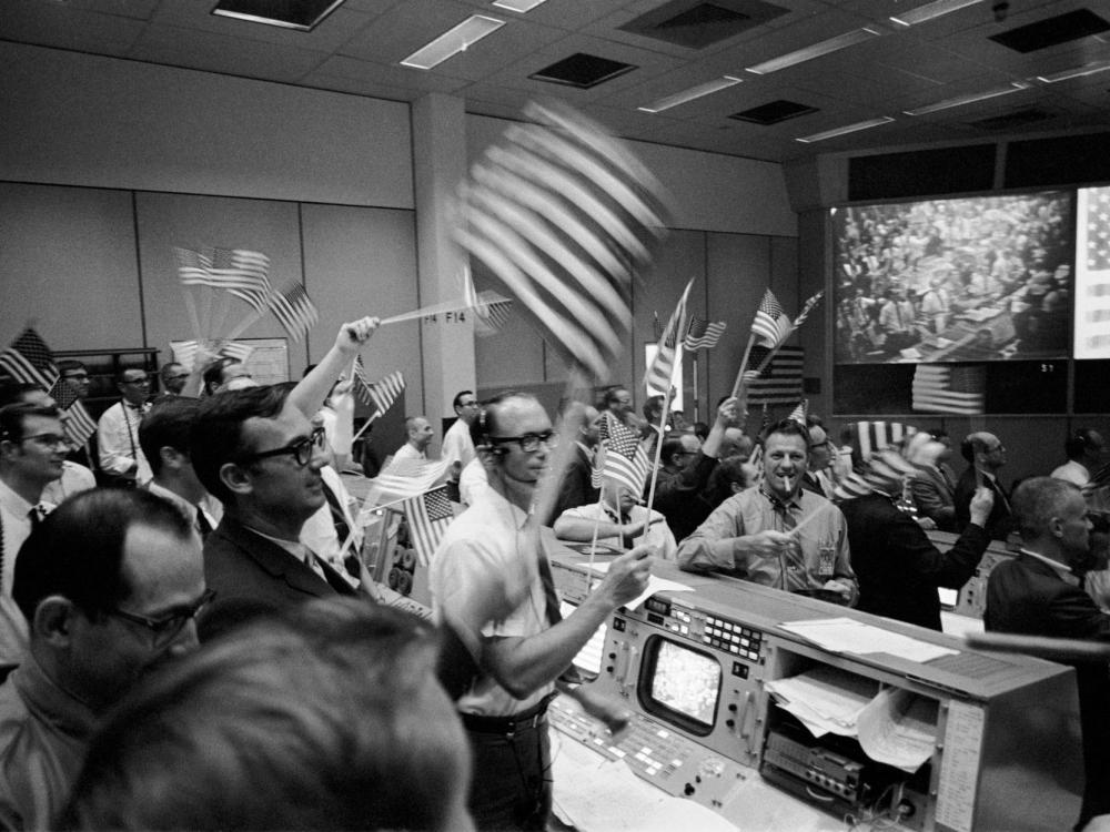 A black and white photograph showing men in business attire standing up from 1960 computer modules and celebrating. They're waving American flags, one is smoking a cigar. On the screens in the front of the room is a large American flag and a field of people. 