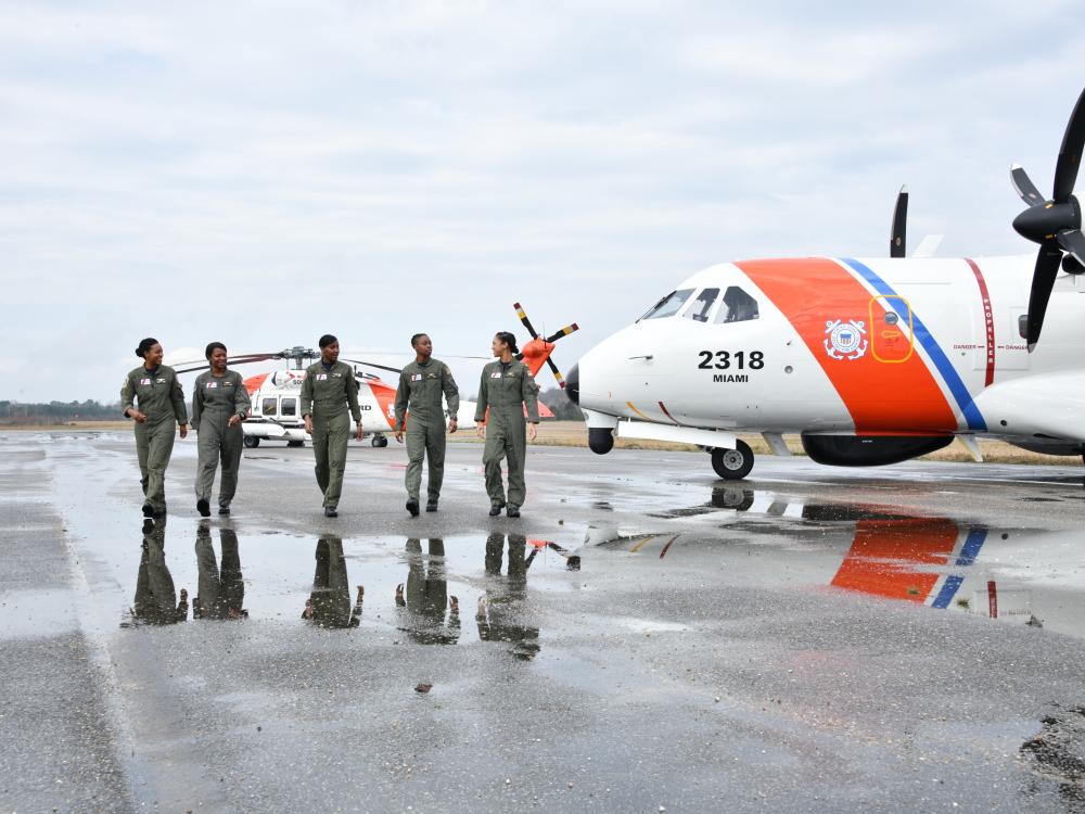 Five black women wearing flight suits walk in front of two Coast Guard helicopters. They are looking at each other, talking and smiling as they wall. 