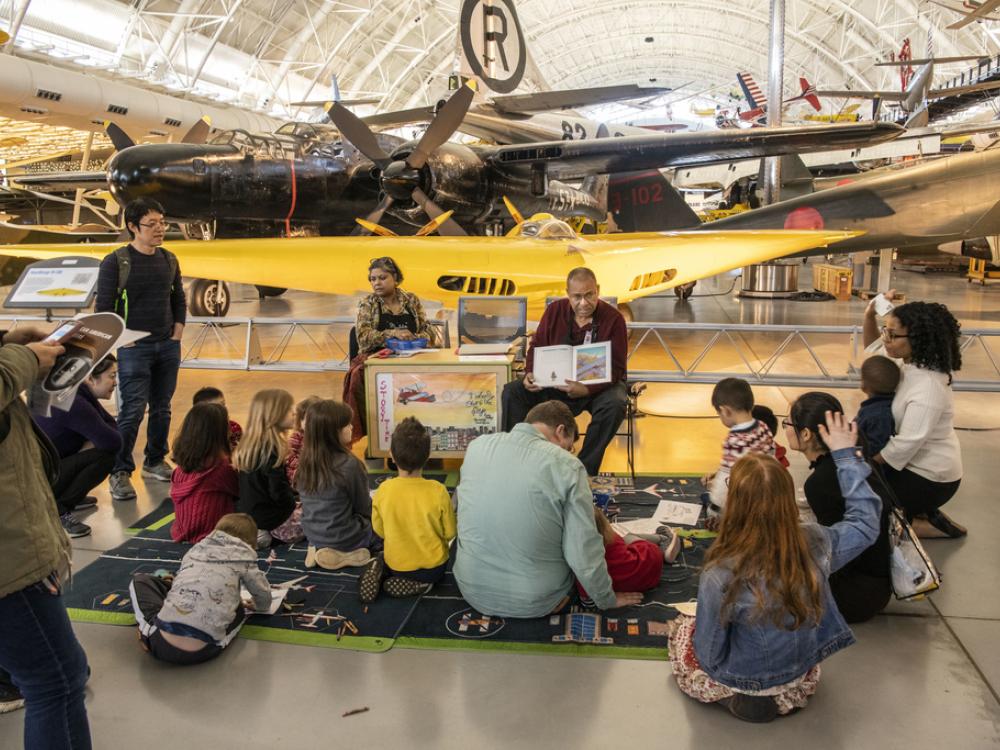 An older couple sits in chairs, reading a children's book, with airplanes in the background. On the ground in front of them are children and parents. 