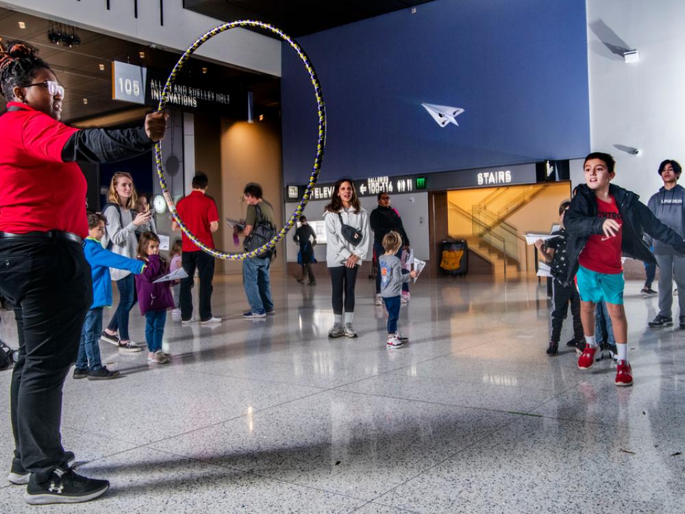 A young Black woman in a red polo holds out a hula hoop. A few feet back a young boy is captured in motion, having just flown a paper airplane. The plane zooms between the two. 