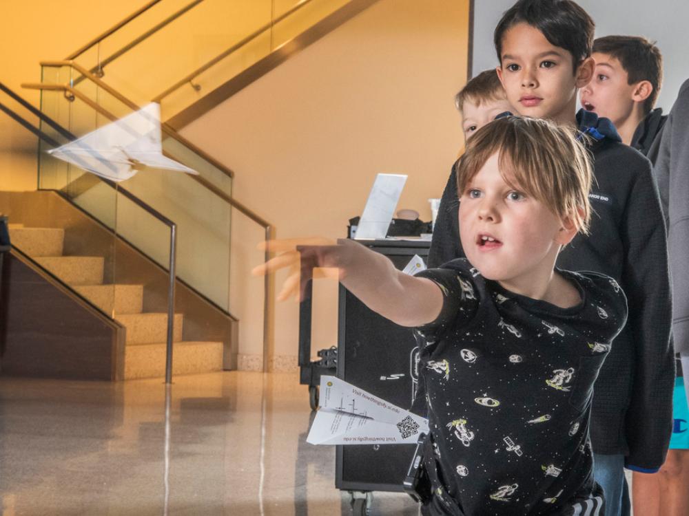 A young visitor is rapt with attention as they throw a paper airplane, with a similar line of young visitors behind them. 