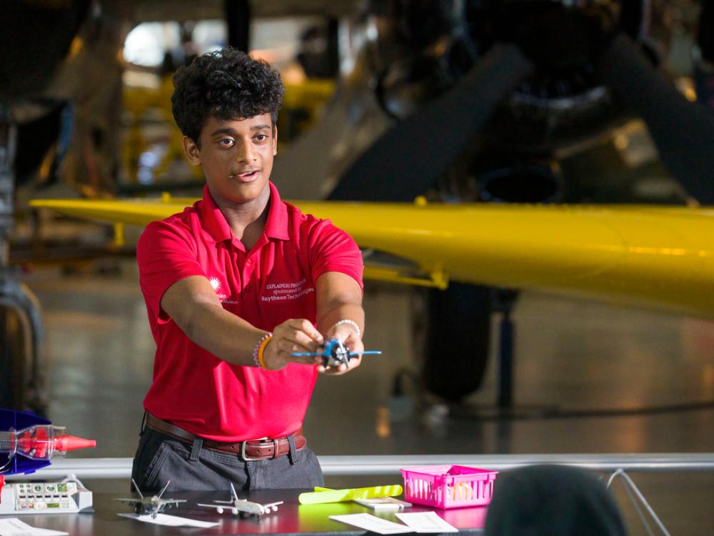 A young man with a light brown skin tone and curly hair wearing a red polo holds out a model of a plane straight in front of him. 