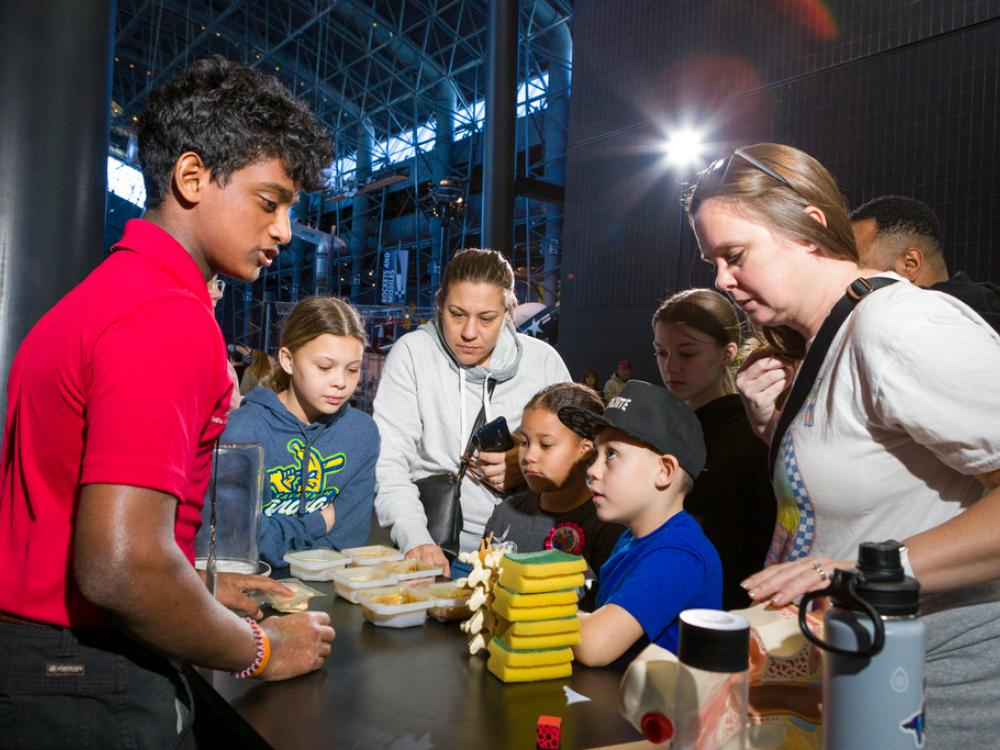 A young man with light brown skin and curly hair wearing a red polo talks to a family with adults and children of all ages, all looking at objects on a cart--except for one young boy who looks up reverently at the Explainer. 