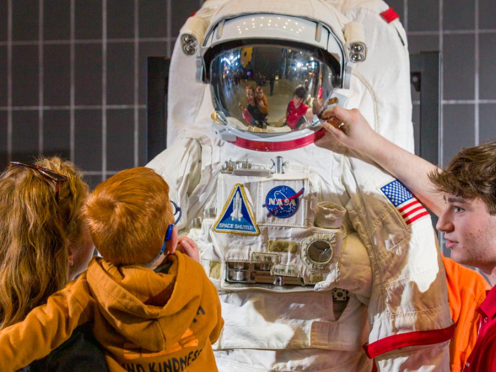 A parent and child look up at a model of a spacesuit, while a young man in a red polo gestures and points. 