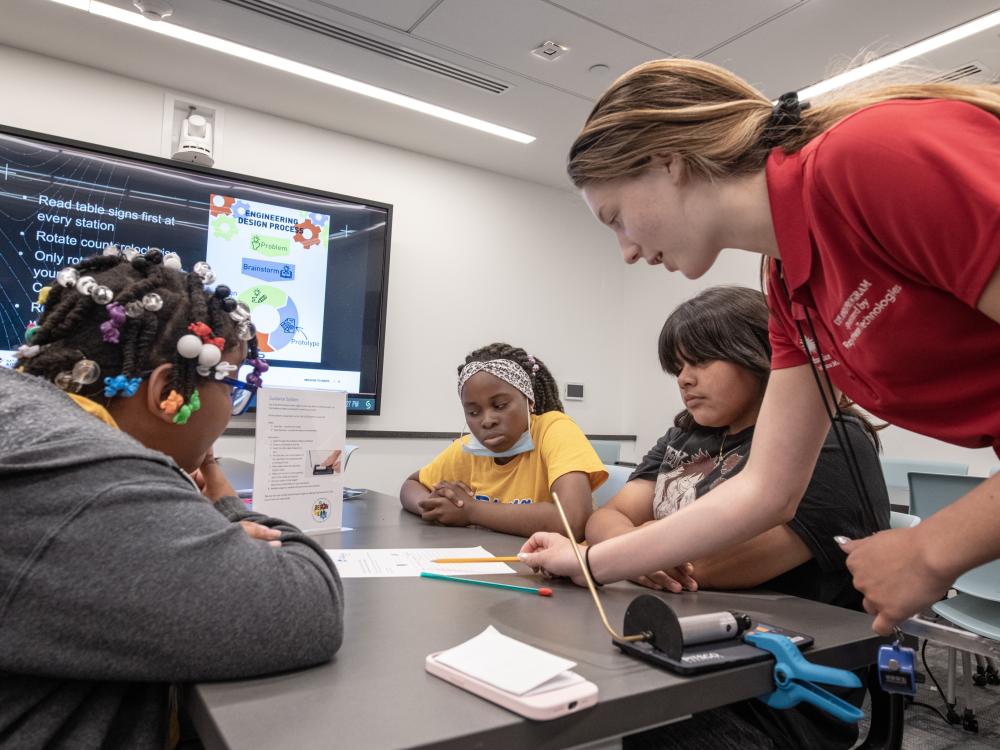 Three teenaged students pay attention as a Museum explainer, a woman in a red polo, explains an activity. 