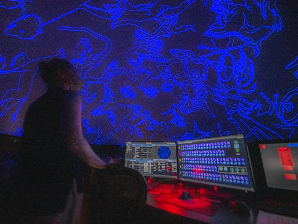 A woman stands at a control booth while images of constellations are projected on a curved planetarium screen. 