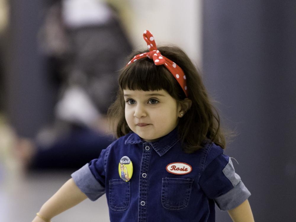 A young girl, approximately 4 years old, struts across the Museum floor in a blue denim jumpsuit and red bandana based on Rosie the Riveter. 