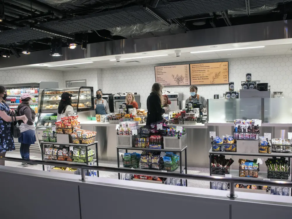  Visitors walk along an aisle in front of a cash register. Food is nearby. 