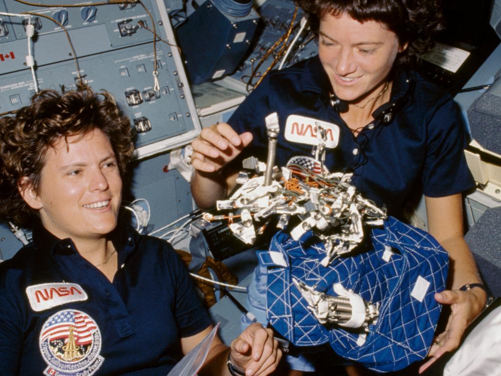 Sally Ride and Kathryn Sullivan, both of whom are white female astronauts, display an object in space.