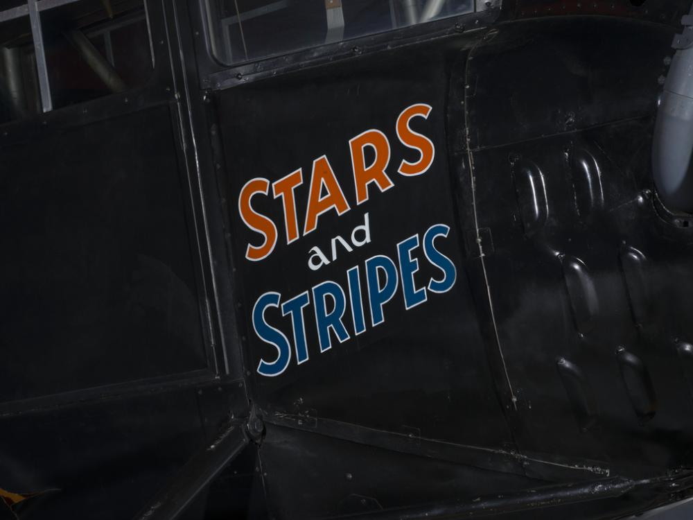 The words Stars and Stripes are painted orange and blue on the side of an airplane. 		