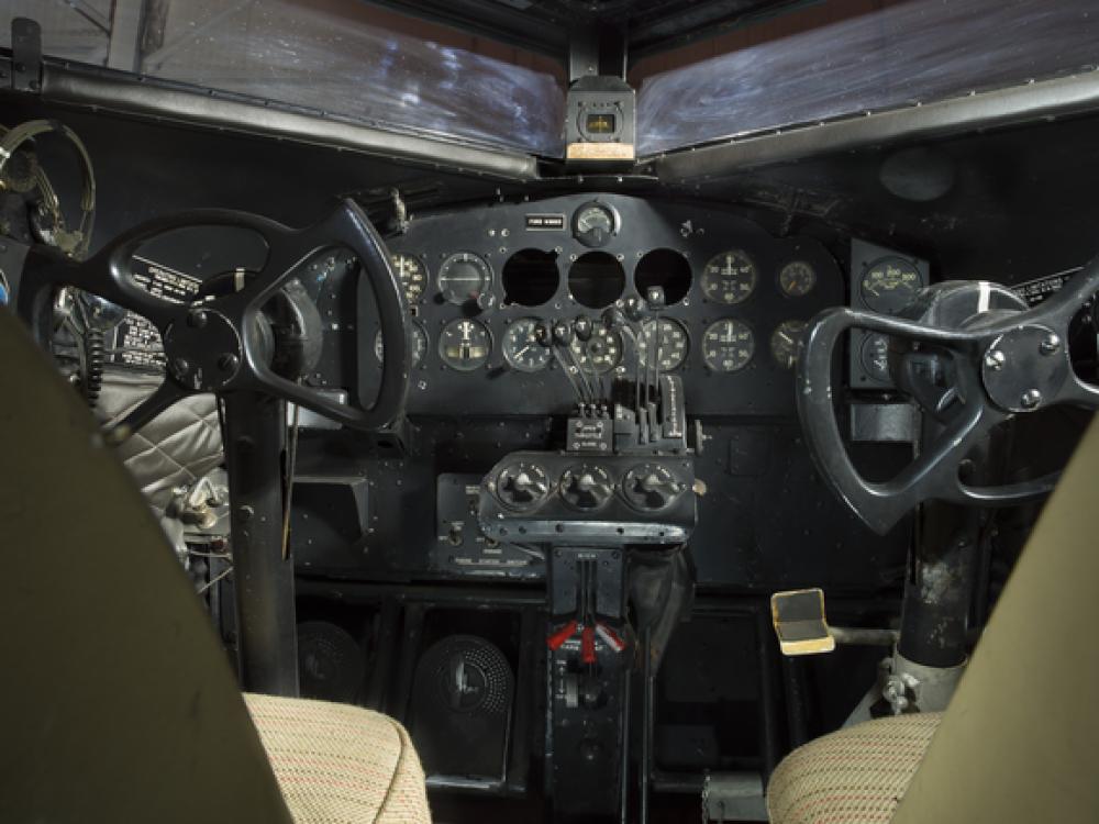 View of the cockpit on the plane. The image was taken behind the seats of the cockpit. The image highlights the instrument panel of the plane. 