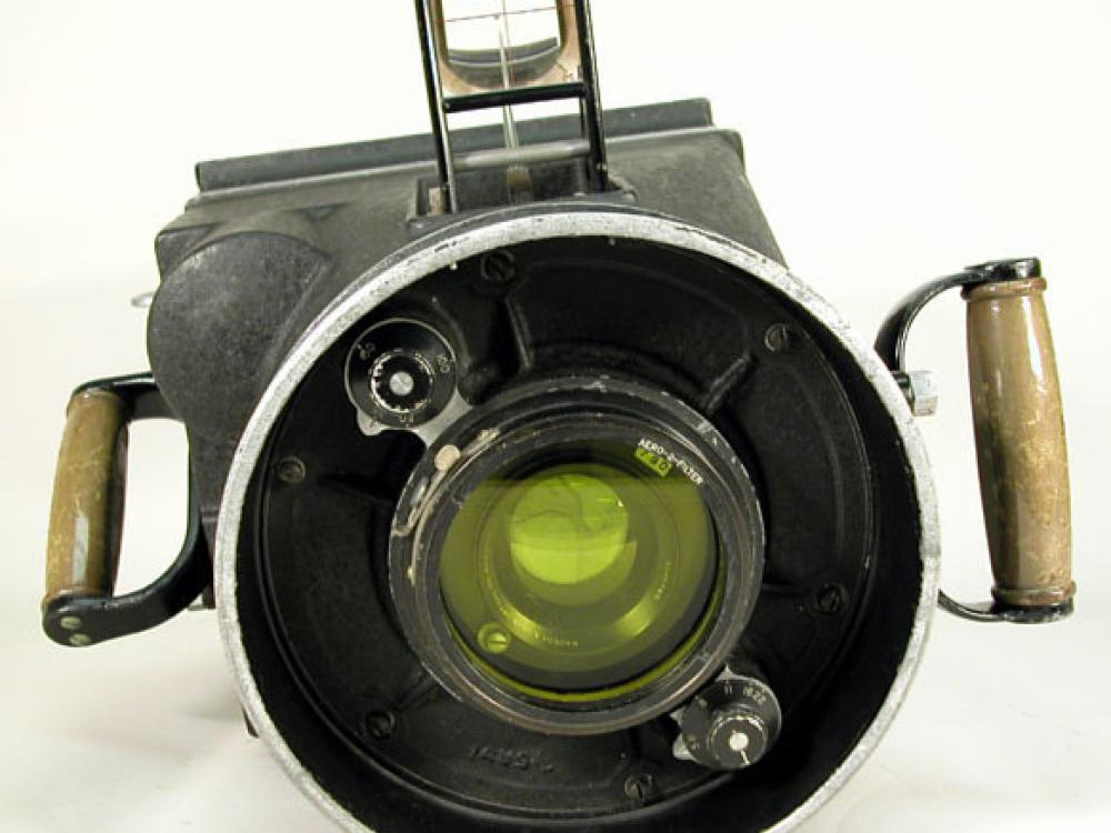 A large circular lens dominates the image. Behind it you can see the body of the camera, from which a viewfinder appears, and handles on either side.