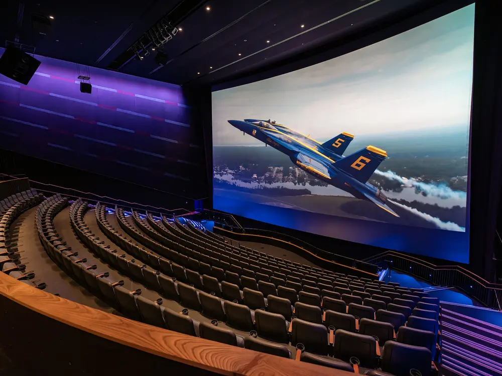 Empty seats face a large screen, stacked in stadium seating. On the screen two blue angels zoom to the upper left hand side of the screen. 