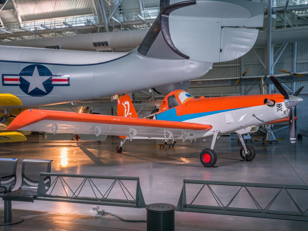 Side view of agricultural plane painted like an orange and white anthropomorphic animated plane, with eyeballs in the cockpit. It sits below a large grey aircraft