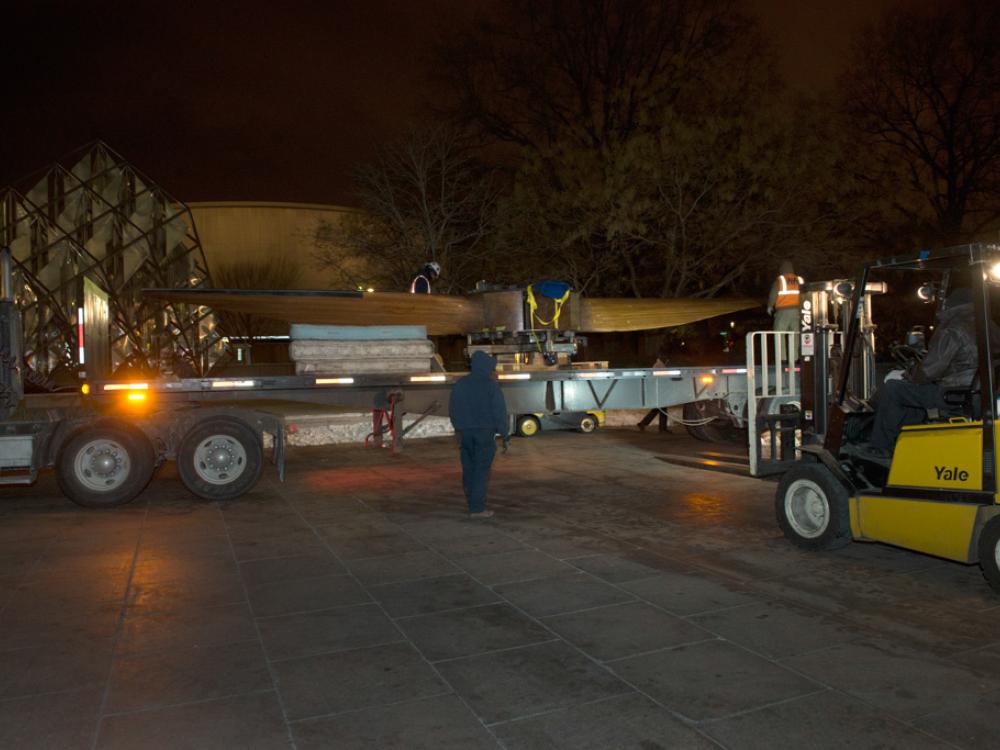 Outside of the Museum, a wind tunnel fan is transported by a truck with a large flatbed into the Museum.