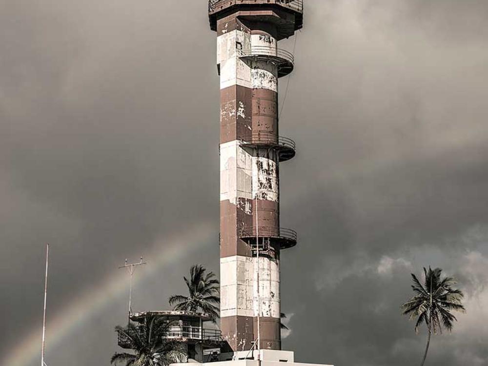 Partial view of an air traffic control tower at Ford Island Airport in Hawaii. The base features an alternating white and brown-colored concrete with three open balconies. Above the base, a single story exists for monitoring aircrafts and a balcony is found throughout the top of the tower.