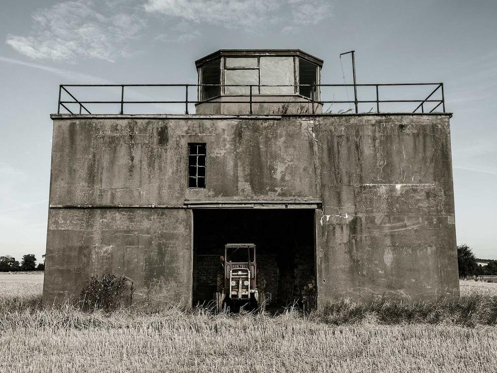 Partial view of the air traffic control tower at East Fortune Field in Scotland. The tower is three stories and features a concrete base with an opening in the center of the first floor. The third floor features at least four different observation angles.