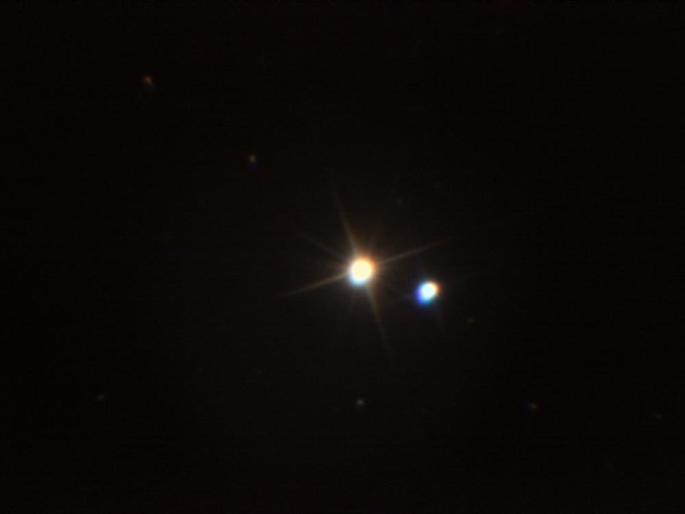 A pair of two stars close together in the night time sky. The left star has a red tint whereas the second, smaller star on the right has a minor blue tint.