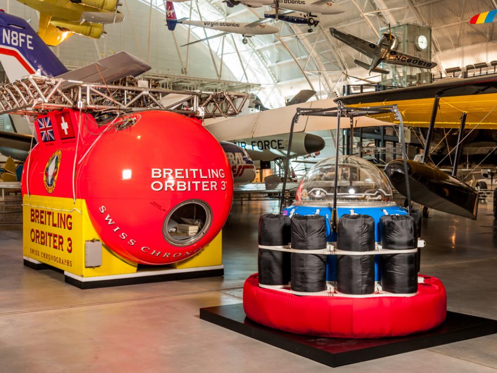 A large red and yellow balloon gondola with a rectangular shape is next to a red and black gondula with a smaller, red circular base and black supports on top of the base. They are both placed in the Museum's Steven F. Udvar-Hazy Center.