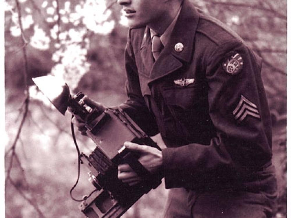 Sgt William E. "Bill" Jones, a white male U.S. Army photographer, crouches as he holds a camera in a forest.