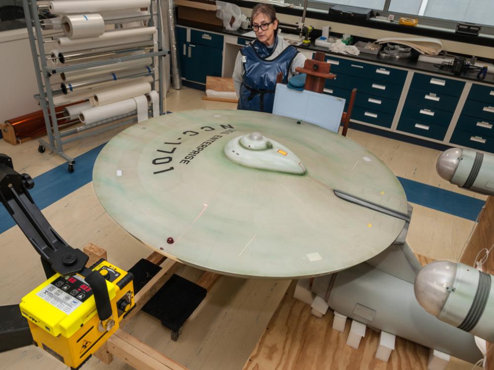 Top view of the starship Enterprise, a gray-colored, dish-shaped starship studio model, with a Smithsonian staff member standing near the studio model. The studio model is undergoing an X-ray scan.