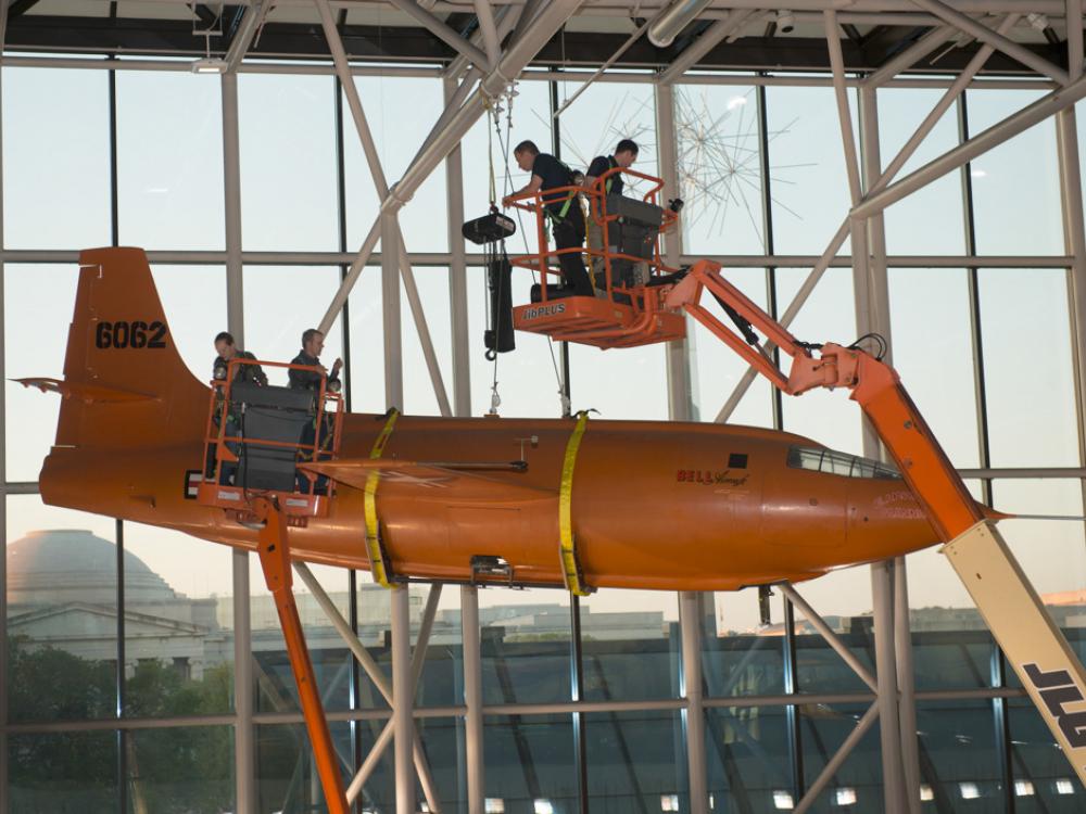 Using two lift machines, Museum employees work to secure the Bell X-1, an orange military monoplane, from the ceiling of the Boeing Milestones of Flight Hall onto the floor.