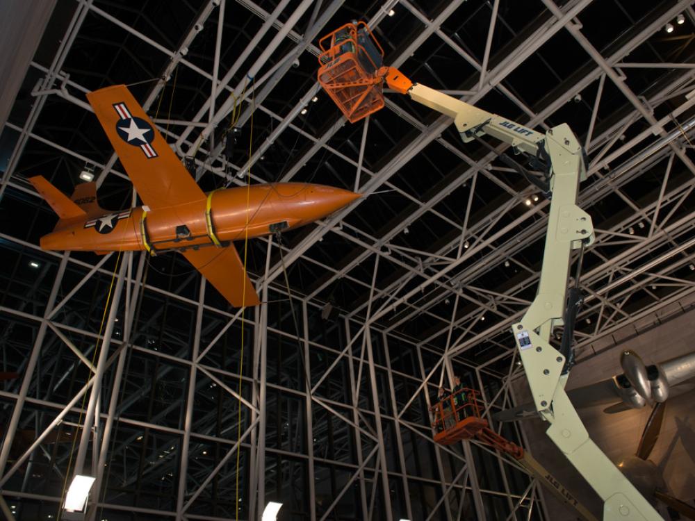 Using a singular lift machine, Museum employees work overnight to lower the Bell X-1, an orange military monoplane, from the ceiling to the floor of the Boeing Milestones of Flight Hall.