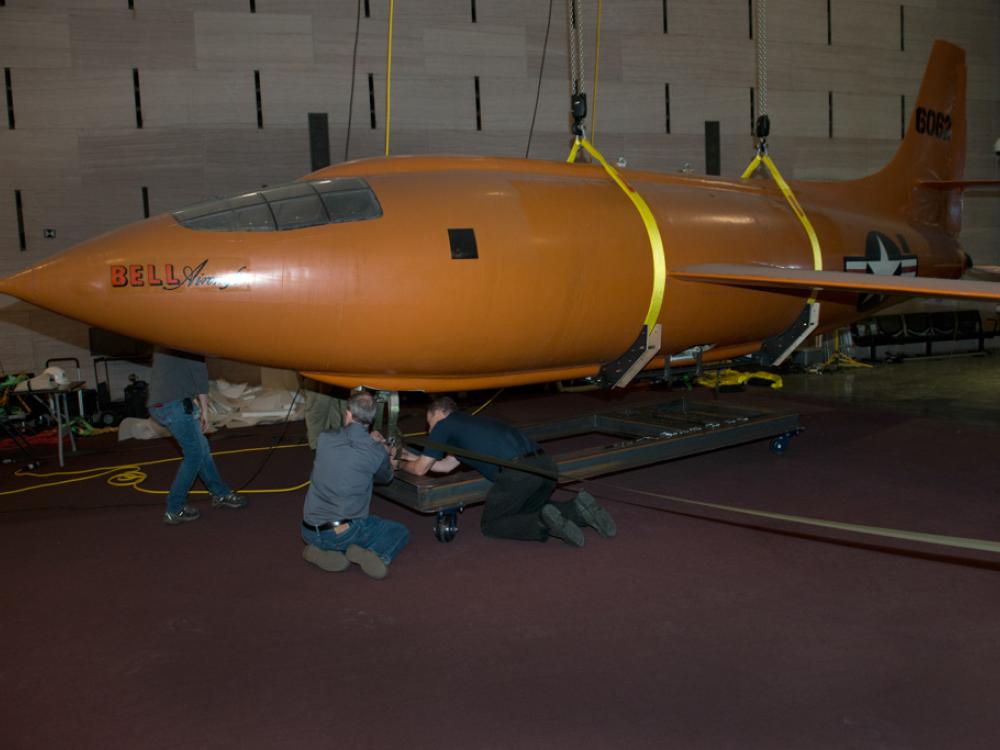 Museum employees secure the fully lowered Bell X-1, an orange military monoplane, as it stands stationary on the floor of the Boeing Milestones of Flight Hall.