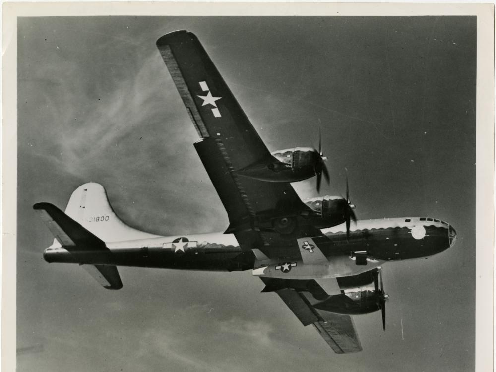 Bottom view of the Bell X-1, a military aircraft, being lifted under a B-29, a larger military aircraft. The B-29 is in flight.