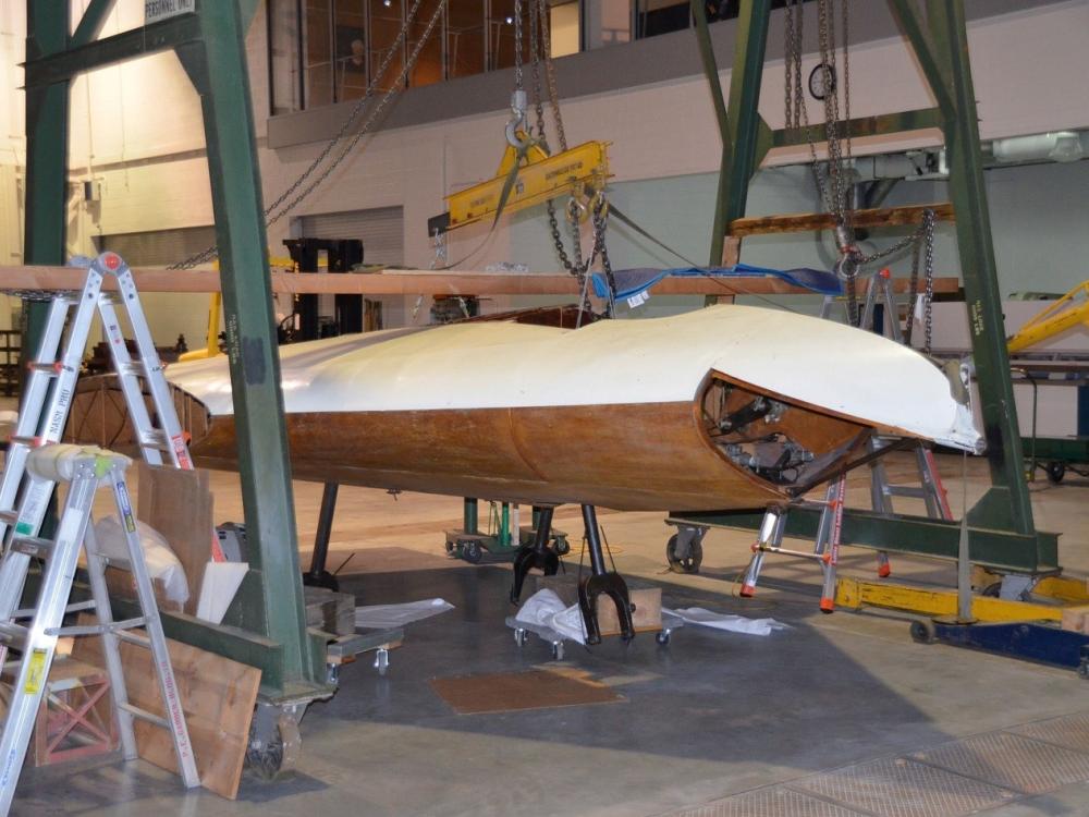 Diagonal side view of part of a white and wooden-colored glider aircraft, which is placed in between two metal frames. A section of clear plastic is removed from the front of the aircraft.