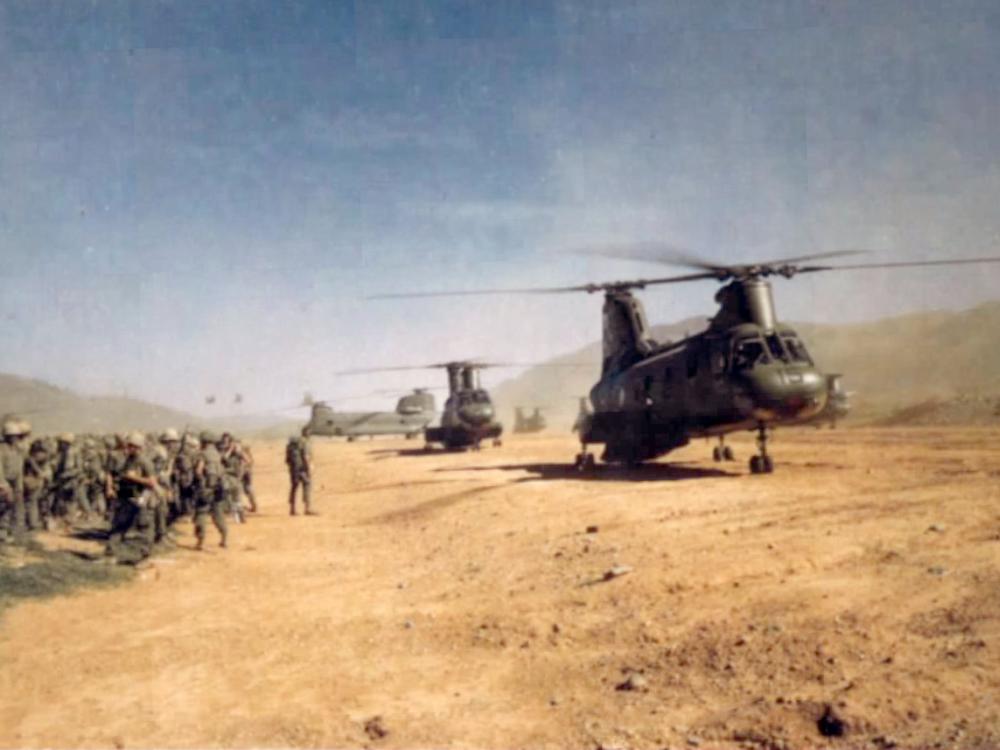 A group of soldiers stand near a sandy plain during the Vietnam War. On the sand, multiple military helicopters are visible and are stationary.