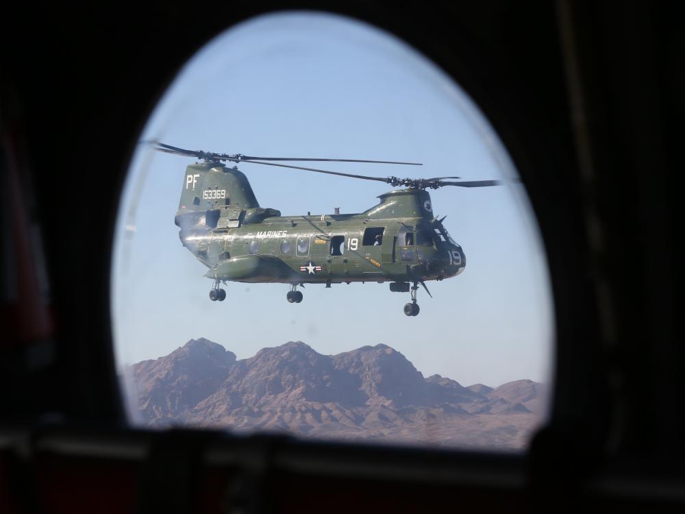 Porthole-style view of green military helicopter with two sets of propellors on top of the helicopter's body. The helicopter is in flight.