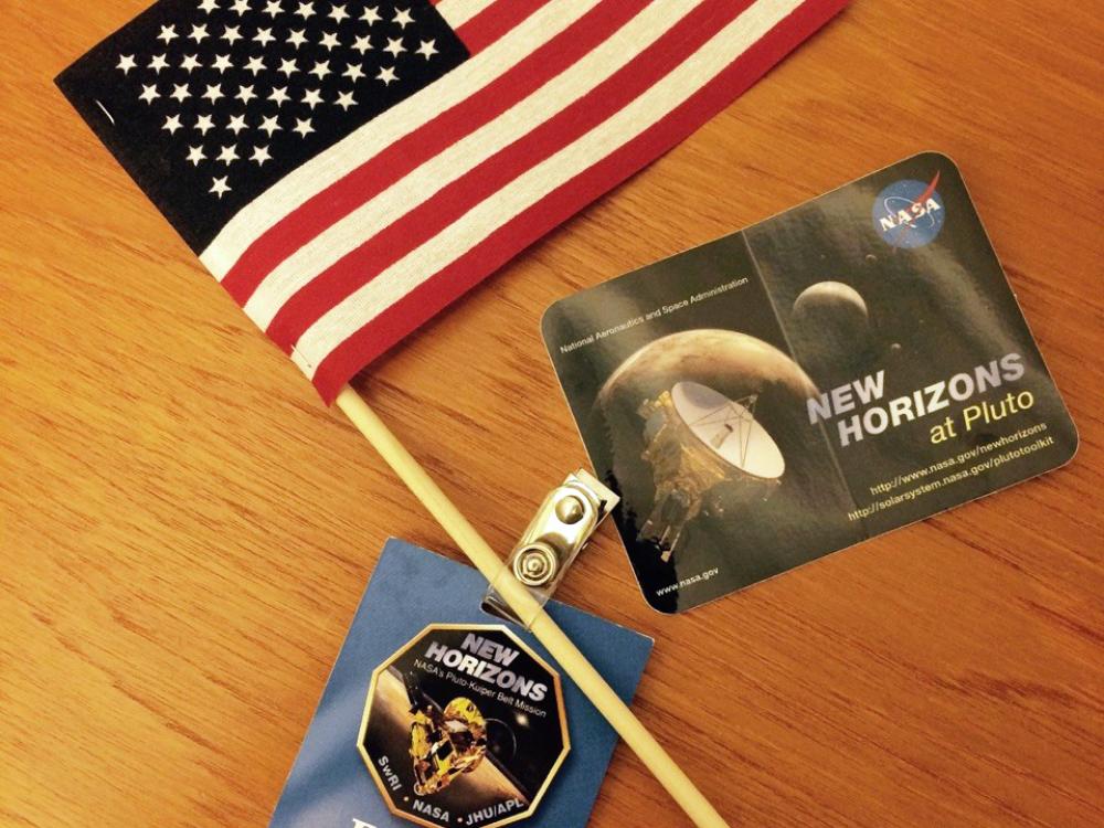 An American flag, blue guest badge, and sticker featuring Pluto that were given out at an event celebrating the New Horizons spacecraft.