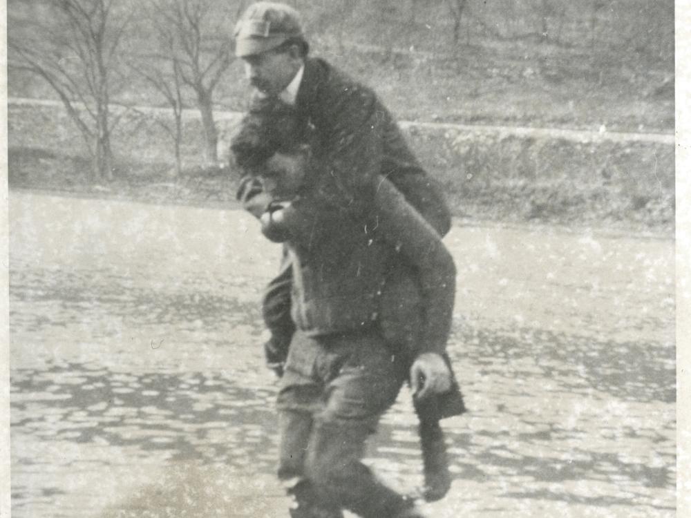 A mechanic working with Orville Wright gives Orville a piggyback as they walk in a river.