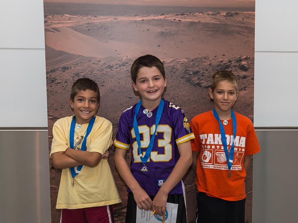 Three boys stand in front of a banner of Mars that is congratulating them on their completion of an astronaut game.