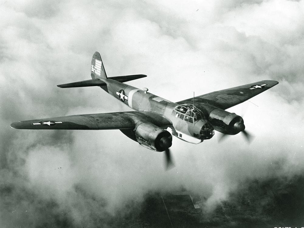 Diagonal front view of military monoplane with twin engines. U.S. star insignia is seen on the wings and rear fuselage. Aircraft is in flight.