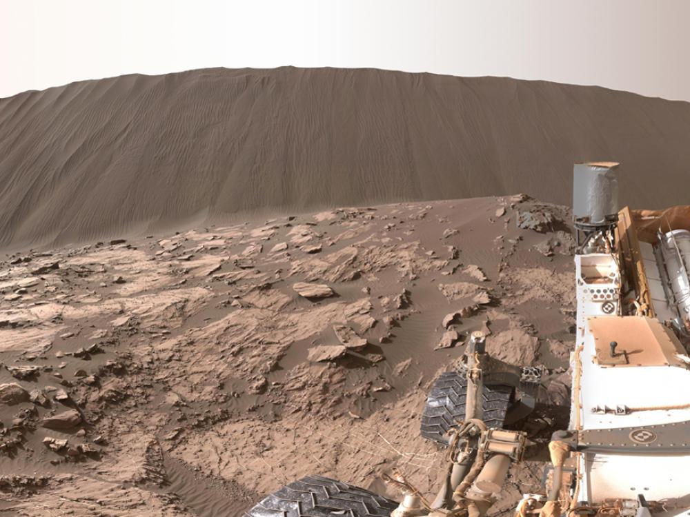 Tall sand dune in the background of a rocky, Mars surface. Curiosity Rover, a robotic surveyor, can be seen on the right side.