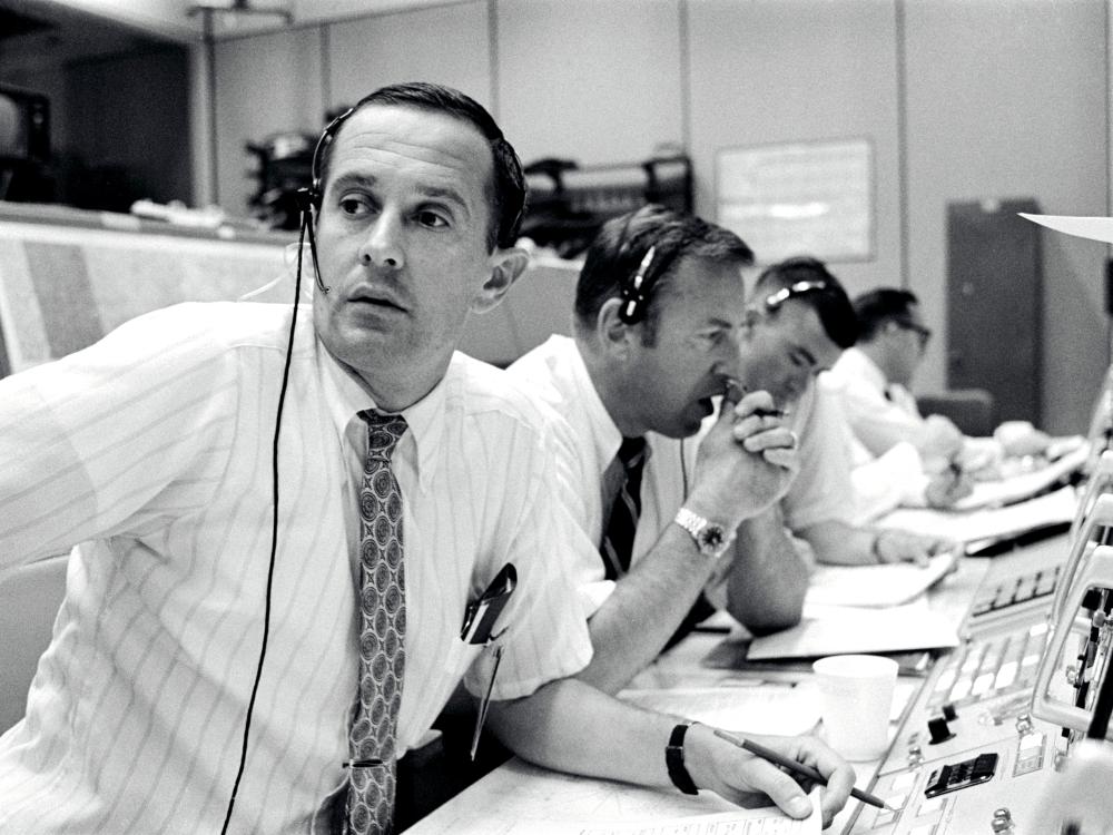 James A. Lovell, a white male NASA astronaut second from the foreground, monitors consoles during the descent of the Apollo 11 lunar module.