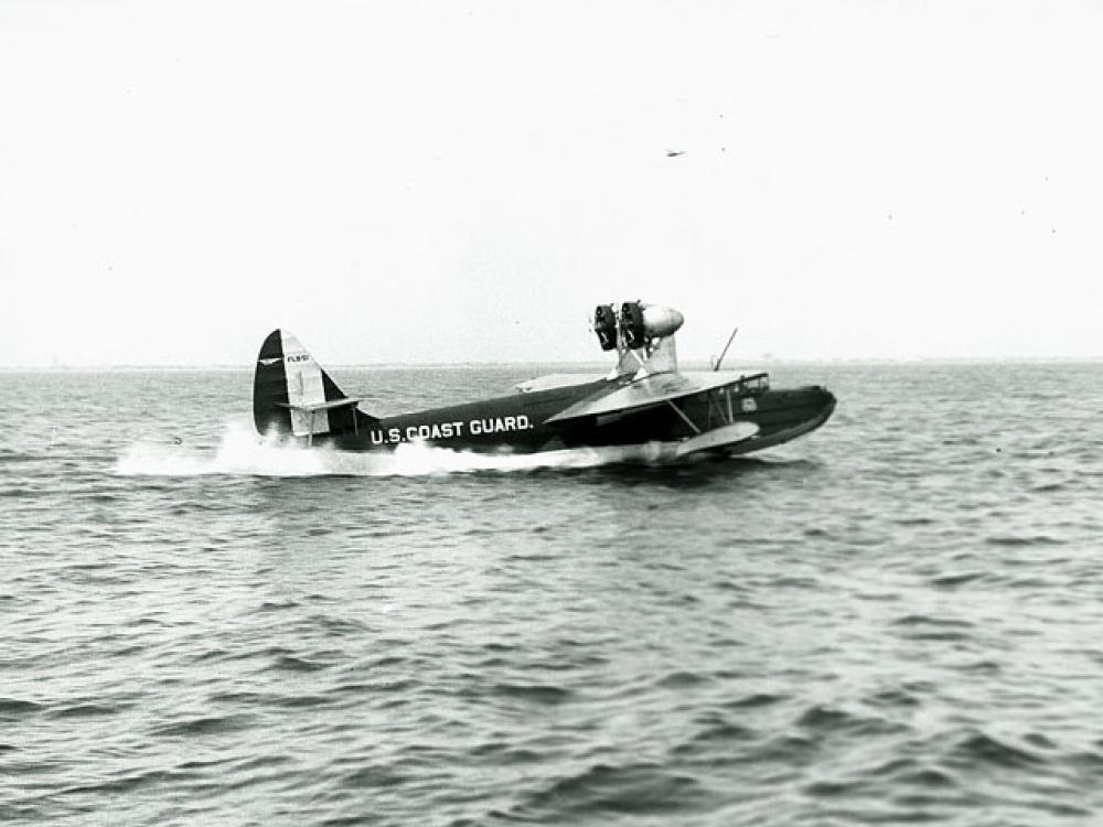 Side view of dark-colored sailplane with the name "U.S. Coast Guard" painted in a lighter color on the back fuselage of the aircraft. Two propellors can be seen on top of the front of the fuselage.