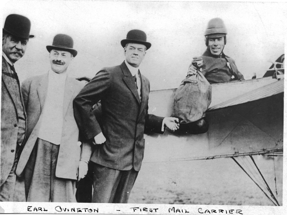 Earle Ovington, a male mail pilot, sits in the cockpit of an aircraft holding a bag of mail.