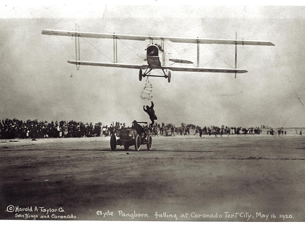 Front view of a biplane during an expedition flight. A person is trying to climb from an automobile below the biplane onto the biplane.