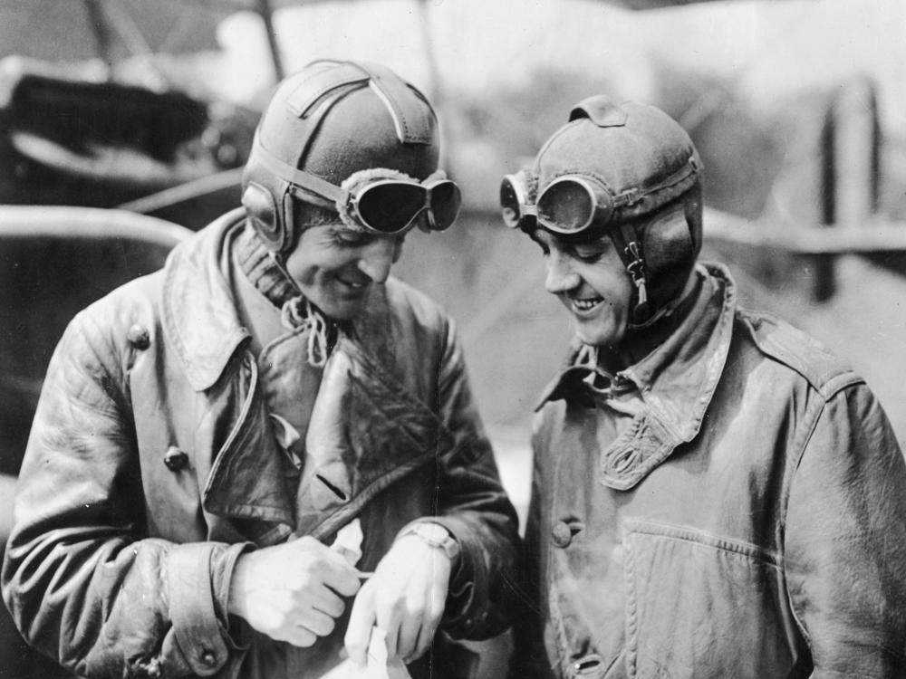 Two male military pilots in aviation gear look at a watch on the hand of the man on the left.