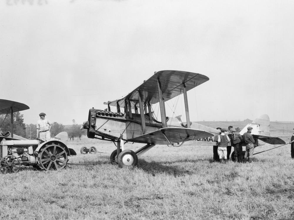 Side view of a light-colored, metal biplane behind a tractor in a field.