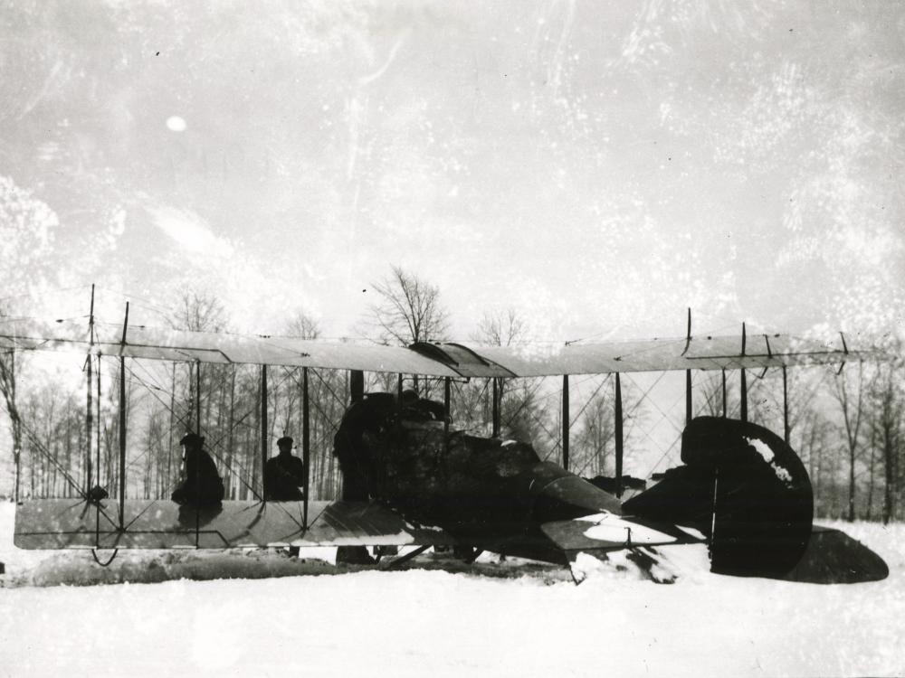 Front view of a biplane partially covered with materials as it stands on snowy ground in a snowstorm.