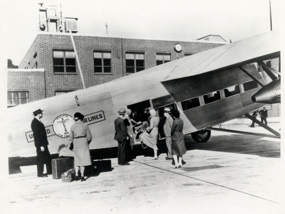 People board a light-colored commercial airplane with the United Air Lines logo on the side of the plane behind where passengers board.