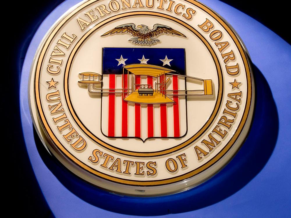 Golden-colored seal of the Civil Aeronautics Board featuring a red, white, and blue flag in the center below a bald eagle. The flag is covered in part by a yellow biplane.