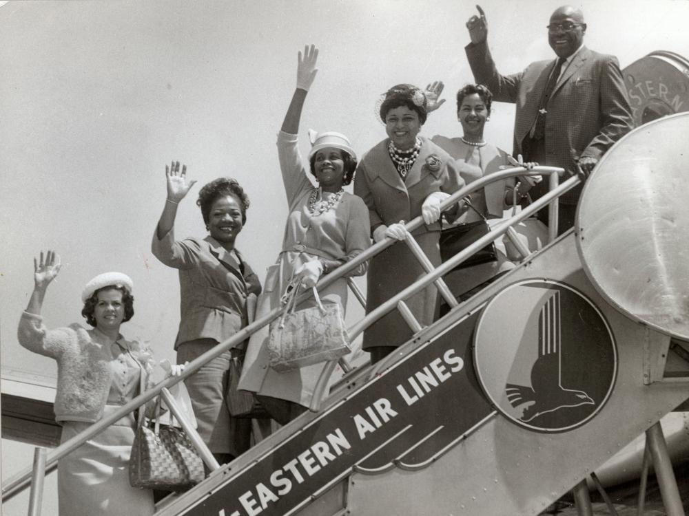 A group of African-American people pose and wave on the boarding steps of an aircraft.